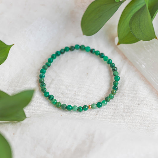 Close-up of an African Jade 4mm gemstone bracelet with a gold accent bead, displayed on a soft white fabric with green leaves surrounding it.