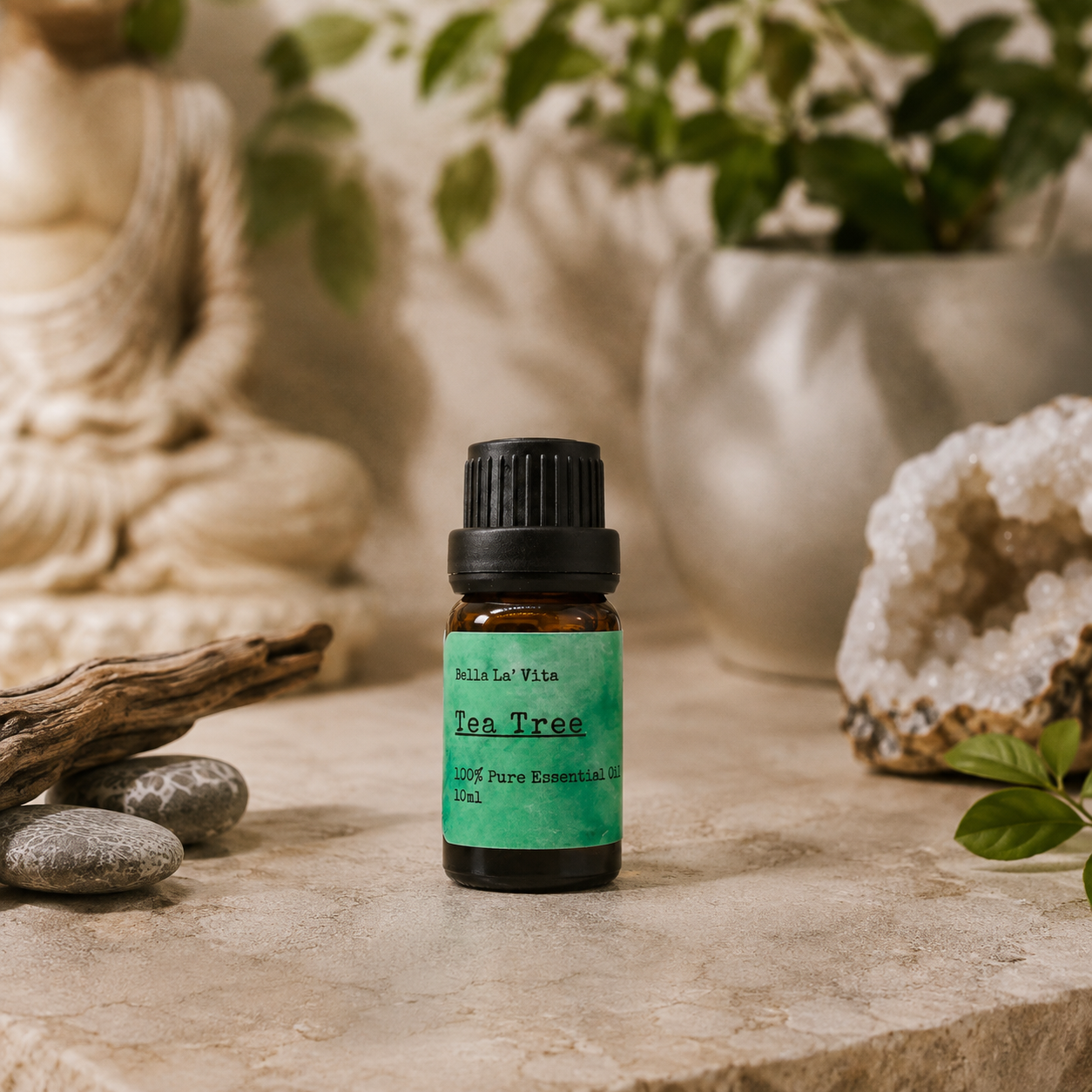Amber 10 ml bottle of Bella La' Vita Tea Tree 100% Pure Essential Oil displayed on a warm zen-inspired stone surface with a Buddha statue, driftwood, smooth stones, greenery, and a crystal geode in the background.