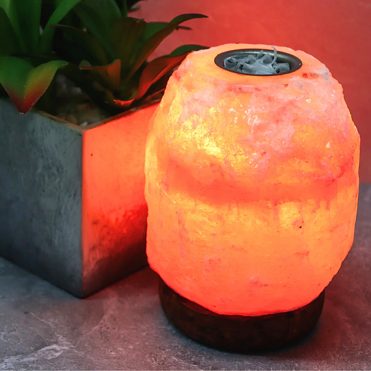 A glowing salt lamp with a rough, textured pinkish-orange body sitting on a dark base, next to a potted green plant on a gray stone surface.