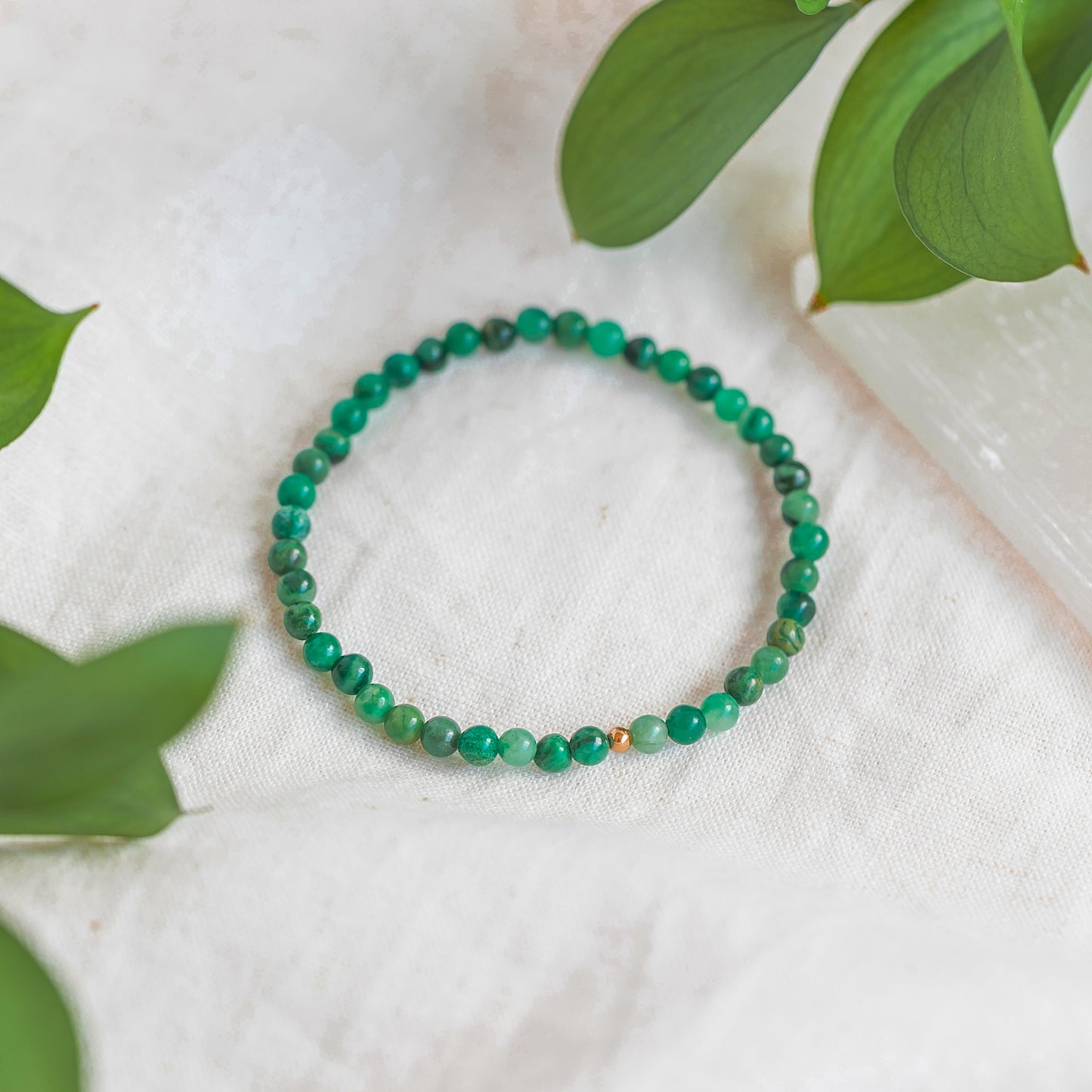 Close-up of an African Jade 4mm gemstone bracelet with a gold accent bead, displayed on a soft white fabric with green leaves surrounding it.