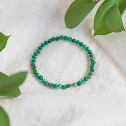 Close-up of an African Jade 4mm gemstone bracelet with a gold accent bead, displayed on a soft white fabric with green leaves surrounding it.