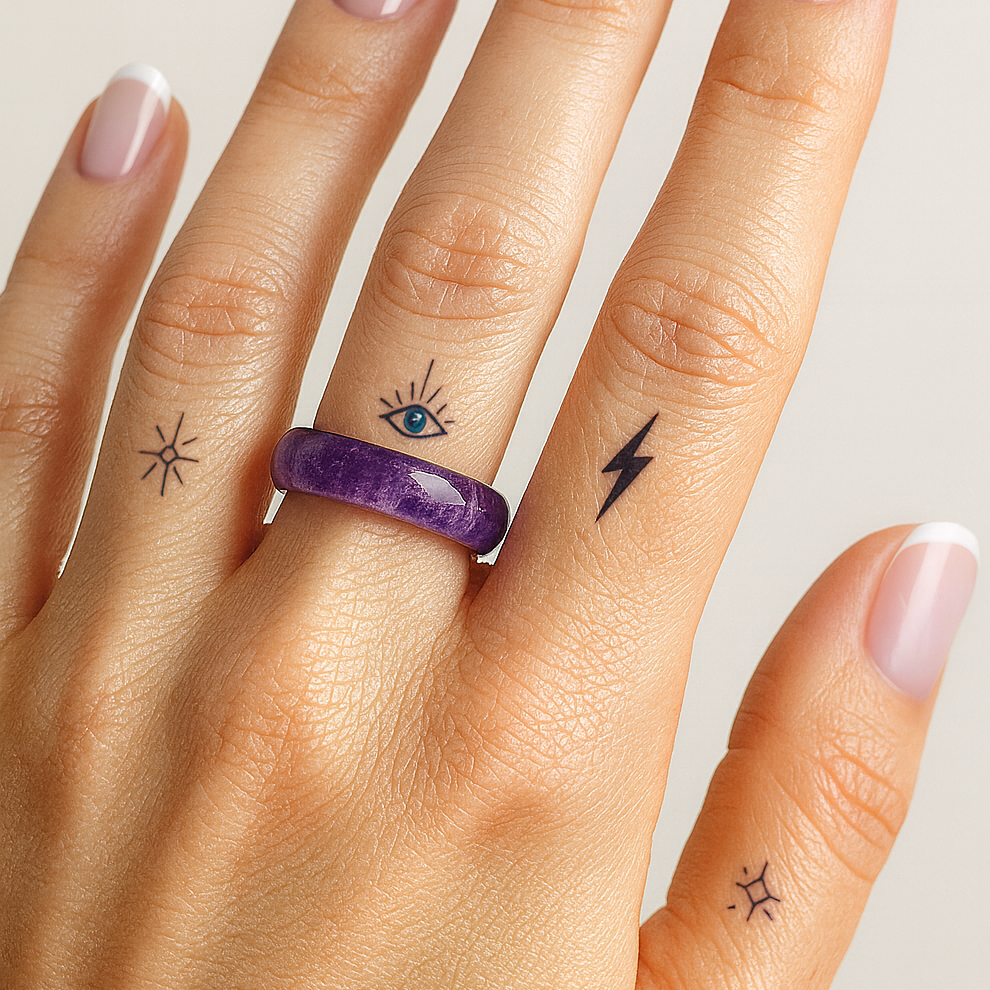 Hand with small tattoos on fingers wearing a purple ring against a neutral background