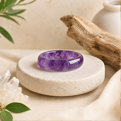 Polished amethyst crystal ring displayed on a textured cream stone pedestal, surrounded by clear quartz, driftwood, green leaves, and soft beige fabric in a warm, zen-inspired setting.