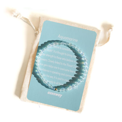Flat-lay of a 4mm Aquamarine beaded bracelet with a gold-filled bead displayed on a blue Azenway meaning card and linen pouch.