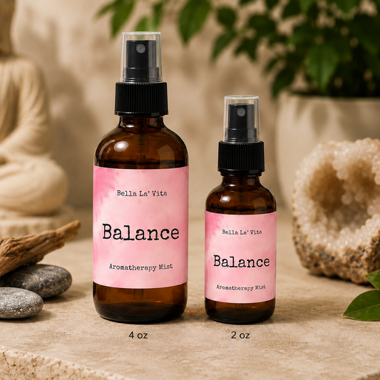 Bella La’ Vita Balance Aromatherapy Mist in 4 oz and 2 oz amber glass spray bottles displayed on a serene, spa-inspired backdrop. The soft pink labels feature the scent name "Balance" in a minimalist design. The bottles are styled with smooth river stones, natural driftwood, a sparkling crystal geode, fresh green leaves, and a softly blurred Buddha statue, creating a peaceful atmosphere that evokes balance, harmony, and everyday wellness.