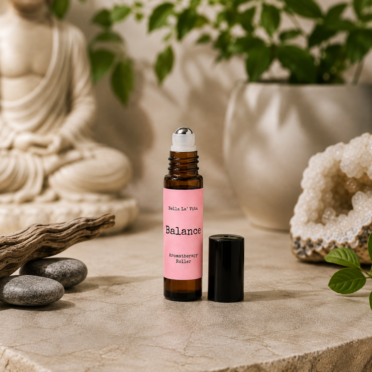 Bella La’ Vita Balance Aromatherapy Roller in an amber glass roller bottle with the cap removed to reveal a stainless steel rollerball applicator. The soft pink label features the scent name "Balance" in a minimalist design. Displayed on a serene, spa-inspired backdrop with smooth river stones, natural driftwood, a sparkling crystal geode, fresh green leaves, and a softly blurred Buddha statue, creating a calming atmosphere that evokes balance, harmony, and everyday wellness.
