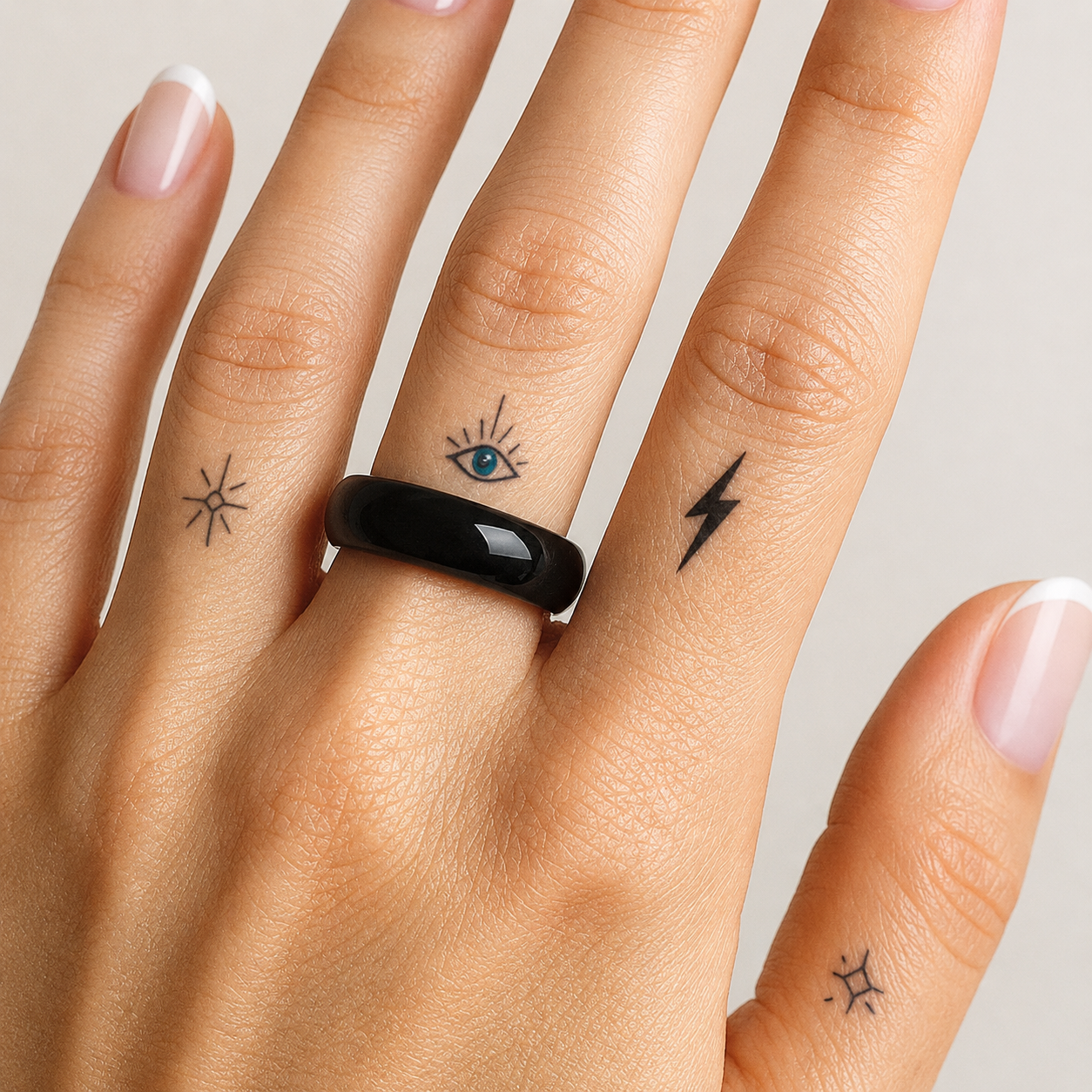 A close-up of a hand wearing a glossy black ring, with small tattoo designs on the fingers, against a light neutral background.