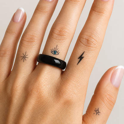 A close-up of a hand wearing a glossy black ring, with small tattoo designs on the fingers, against a light neutral background.