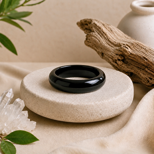 Polished black obsidian crystal ring with a deep glossy finish displayed on a textured cream stone pedestal, surrounded by clear quartz, driftwood, greenery, and soft beige fabric.