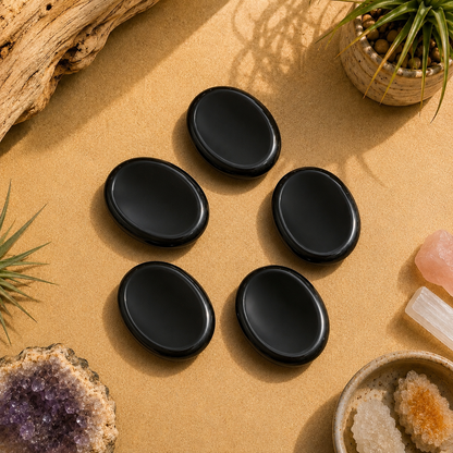 Five polished Black Obsidian worry stones displayed on a warm neutral surface with natural wood, crystals, and botanical accents. Smooth oval palm stones for grounding, meditation, stress relief, mindfulness, and crystal healing.