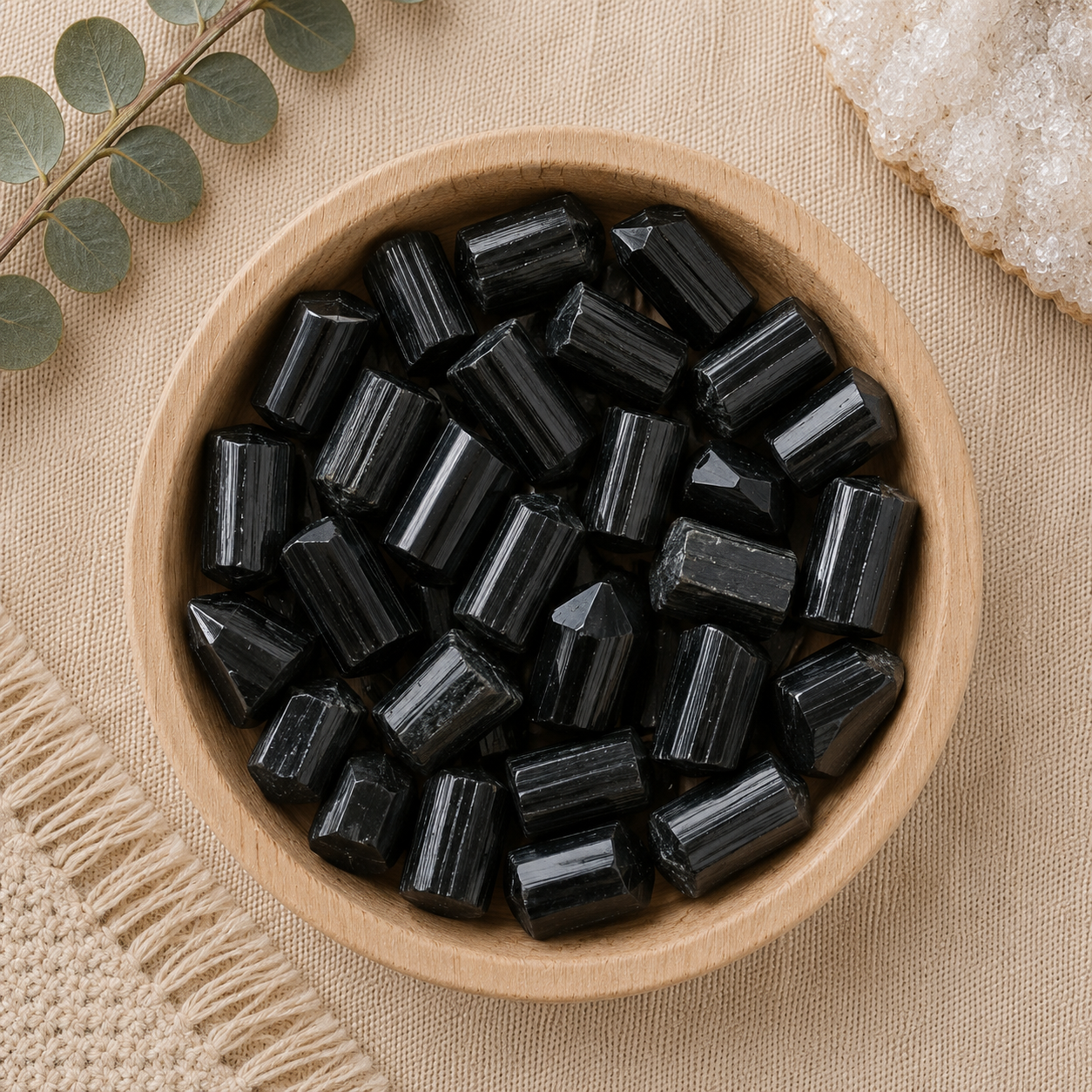 Natural black tourmaline crystal points displayed in a round bamboo bowl on a soft beige linen background. The polished crystals feature their signature vertical striations and pointed terminations, while eucalyptus leaves, a woven cream textile, and a white quartz cluster create a warm, minimalist, zen-inspired flat lay.