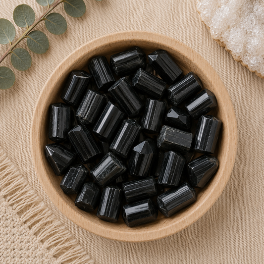 Natural black tourmaline crystal points displayed in a round bamboo bowl on a soft beige linen background. The polished crystals feature their signature vertical striations and pointed terminations, while eucalyptus leaves, a woven cream textile, and a white quartz cluster create a warm, minimalist, zen-inspired flat lay.