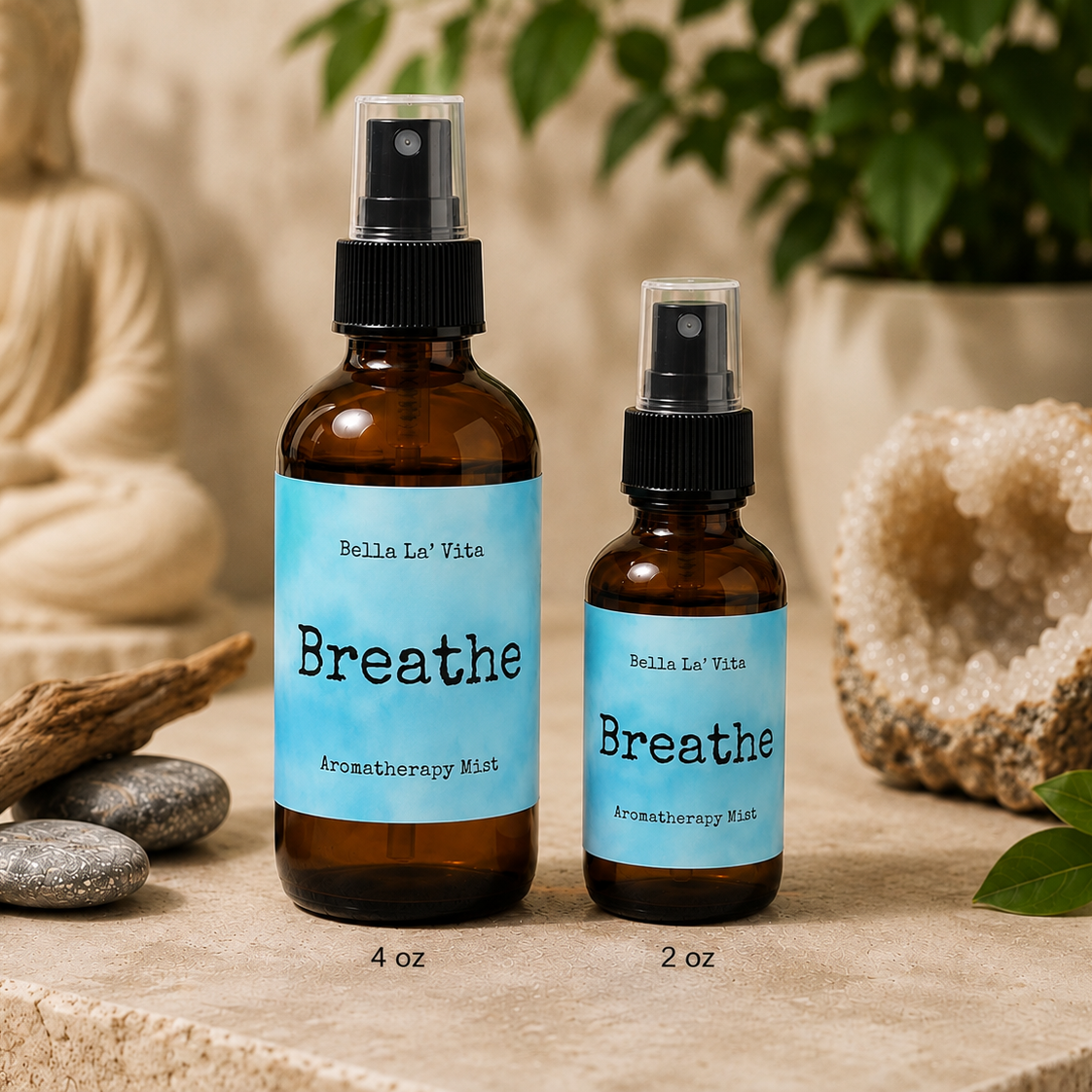 Bella La’ Vita Breathe Aromatherapy Mist in 4 oz and 2 oz amber glass spray bottles displayed on a serene, spa-inspired backdrop. The soft blue labels feature the scent name "Breathe" in a minimalist design. The bottles are styled with smooth river stones, natural driftwood, a sparkling crystal geode, fresh green leaves, and a softly blurred Buddha statue, creating a fresh, calming atmosphere that inspires deep breaths and everyday relaxation.
