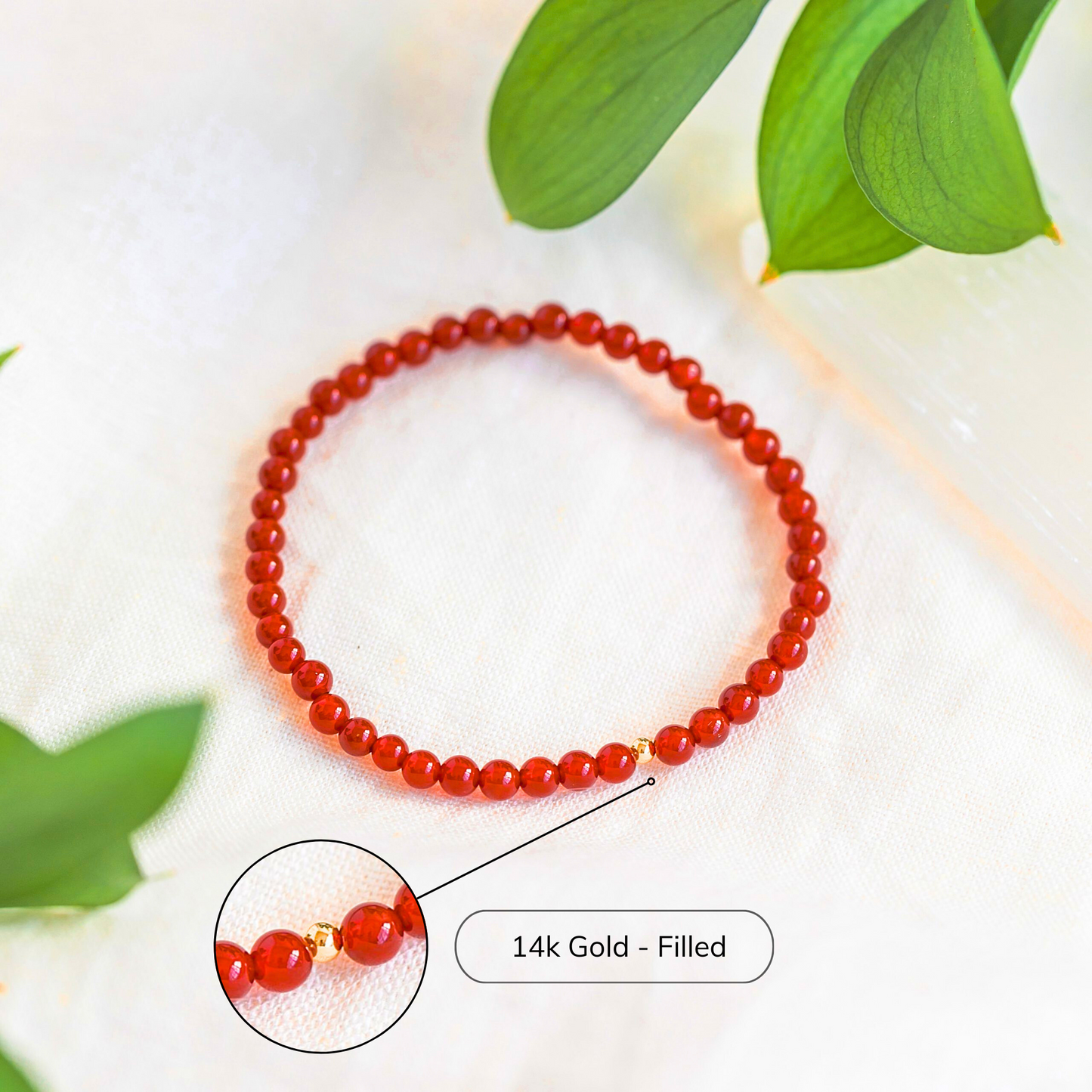 A close-up image of a red carnelian gemstone bracelet with a single 14k gold-filled bead, styled on a soft white background with greenery.
