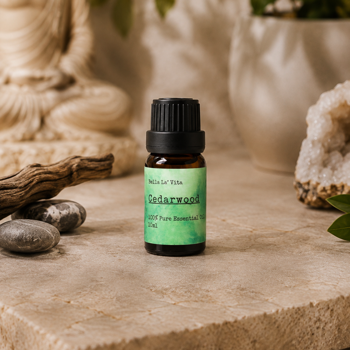 Amber 10 ml bottle of Bella La' Vita Cedarwood 100% Pure Essential Oil displayed on a warm zen-inspired stone surface with a Buddha statue, driftwood, smooth stones, greenery, and a crystal geode in the background.