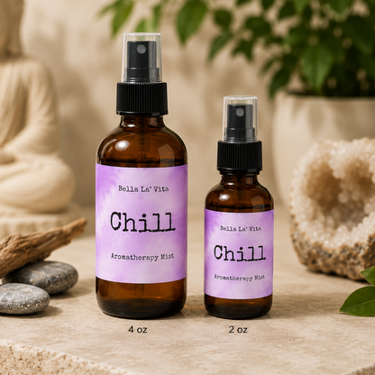 Bella La’ Vita Chill Aromatherapy Mist in 4 oz and 2 oz amber glass spray bottles displayed on a serene spa-inspired backdrop. The lavender watercolor labels feature the scent name "Chill" with a minimalist design. The bottles are styled with smooth river stones, driftwood, a white crystal geode, green leaves, and a softly blurred Buddha statue, creating a calming, zen atmosphere ideal for relaxation and aromatherapy.
