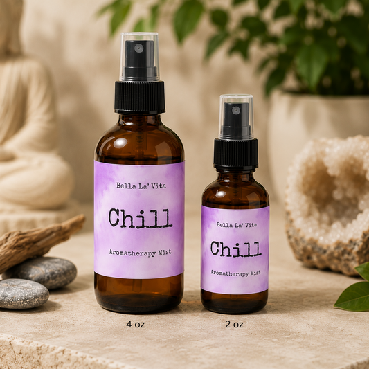Bella La’ Vita Chill Aromatherapy Mist in 4 oz and 2 oz amber glass spray bottles displayed on a serene spa-inspired backdrop. The lavender watercolor labels feature the scent name "Chill" with a minimalist design. The bottles are styled with smooth river stones, driftwood, a white crystal geode, green leaves, and a softly blurred Buddha statue, creating a calming, zen atmosphere ideal for relaxation and aromatherapy.