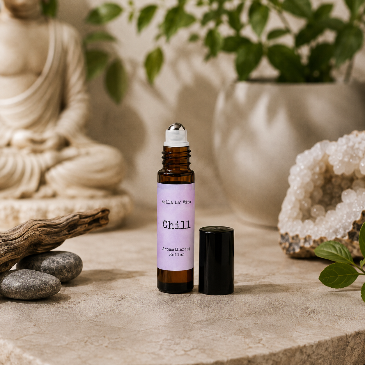 Bella La’ Vita Chill Aromatherapy Roller in an amber glass roll-on bottle with the cap removed to reveal a stainless steel rollerball applicator. The soft lavender label features the scent name "Chill" in a minimalist design. Displayed on a serene, spa-inspired backdrop with smooth river stones, natural driftwood, a sparkling crystal geode, fresh green leaves, and a softly blurred Buddha statue, creating a peaceful atmosphere that evokes relaxation and everyday wellness.