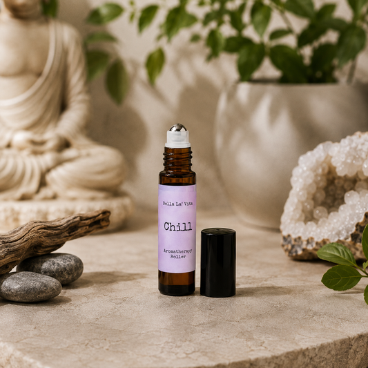 Bella La’ Vita Chill Aromatherapy Roller in an amber glass roll-on bottle with the cap removed to reveal a stainless steel rollerball applicator. The soft lavender label features the scent name "Chill" in a minimalist design. Displayed on a serene, spa-inspired backdrop with smooth river stones, natural driftwood, a sparkling crystal geode, fresh green leaves, and a softly blurred Buddha statue, creating a peaceful atmosphere that evokes relaxation and everyday wellness.