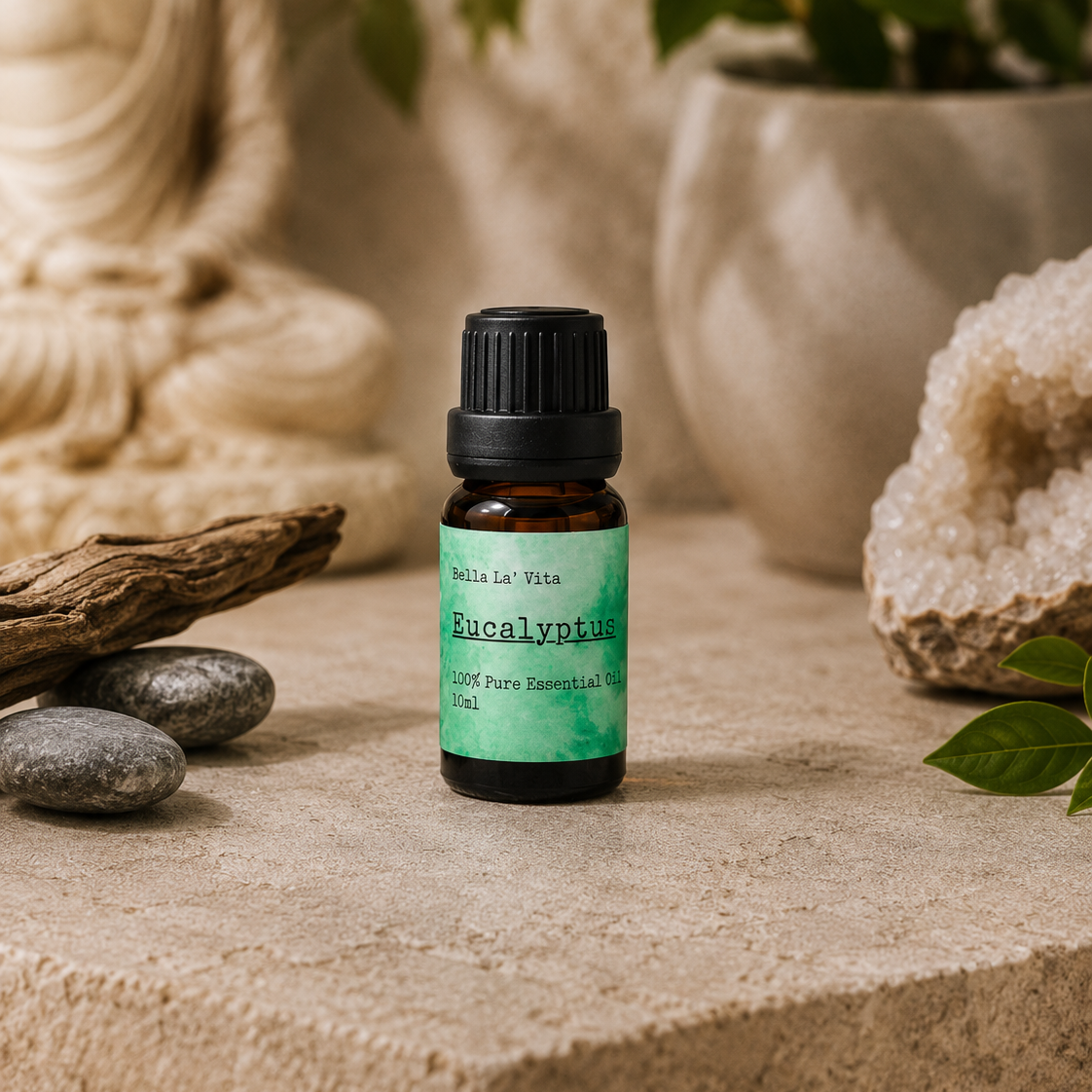 Amber 10 ml bottle of Bella La' Vita Eucalyptus 100% Pure Essential Oil displayed on a warm zen-inspired stone surface with a Buddha statue, driftwood, smooth stones, greenery, and a crystal geode in the background.