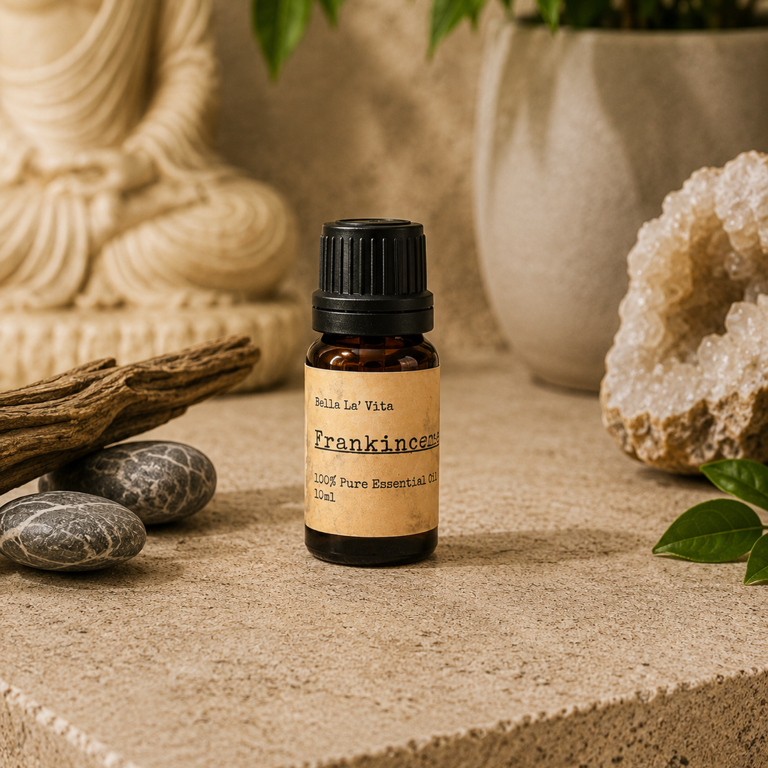 Amber 10 ml bottle of Bella La' Vita Patchouli 100% Pure Essential Oil displayed on a warm zen-inspired stone surface with a Buddha statue, driftwood, smooth stones, greenery, and a crystal geode in the background.