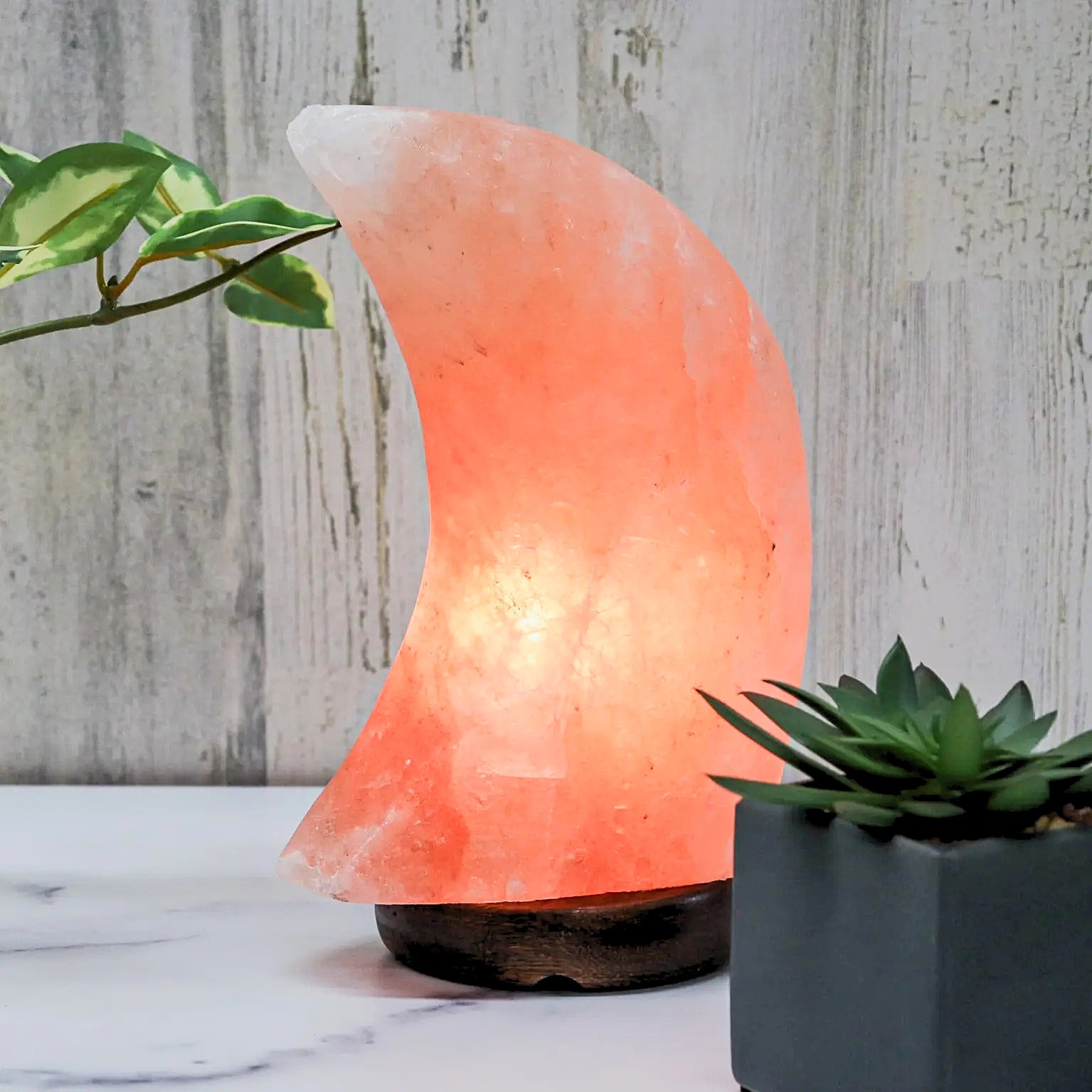 A crescent-shaped Himalayan salt lamp glowing with warm orange light on a dark wooden base, styled on a white marble surface with potted green plants against a light gray wood-plank wall.