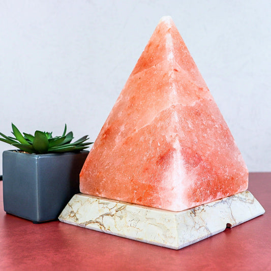 A pink Himalayan salt lamp shaped like a pyramid, resting on a white marble base, with a small potted succulent to its left on a red surface against a light gray background.