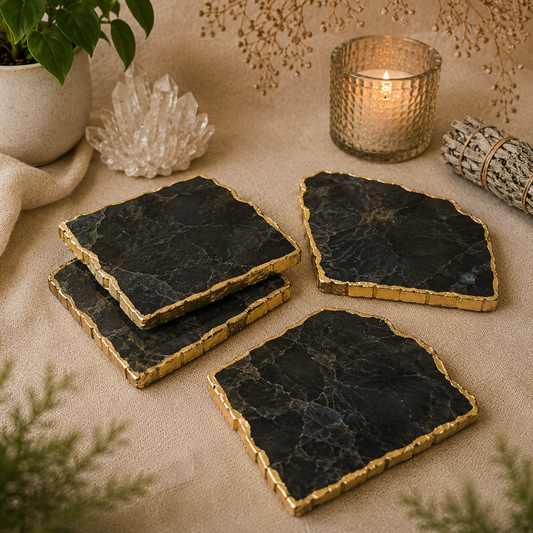 Set of four handcrafted black stone crystal coasters with elegant gold edges displayed on soft beige linen. The natural coasters are styled beside a lit candle, quartz crystal cluster, white sage bundle, greenery, and dried botanicals in a warm, calming boho-inspired setting.