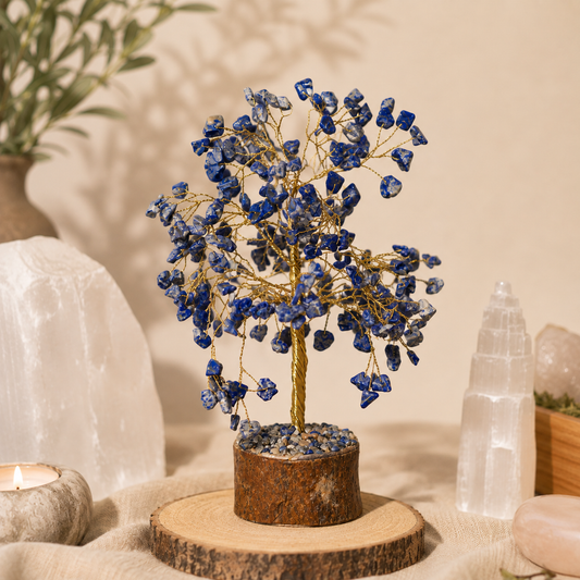 Handcrafted Lapis Lazuli crystal tree featuring deep blue gemstone leaves with natural gold flecks on gold wire branches and a natural wood trunk, displayed on a rustic wood slice. Styled with selenite, Himalayan salt, rose quartz, a candle, and earthy décor in a warm, calming boho-inspired setting.