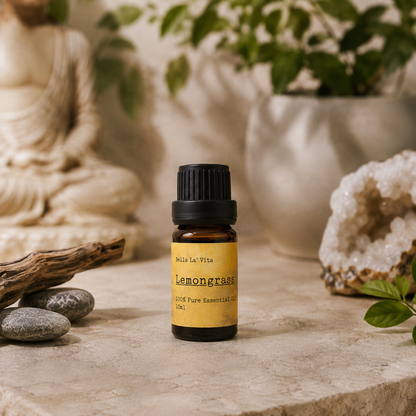 Amber 10 ml bottle of Bella La' Vita Lemongrass 100% Pure Essential Oil displayed on a warm zen-inspired stone surface with a Buddha statue, driftwood, smooth stones, greenery, and a crystal geode in the background.