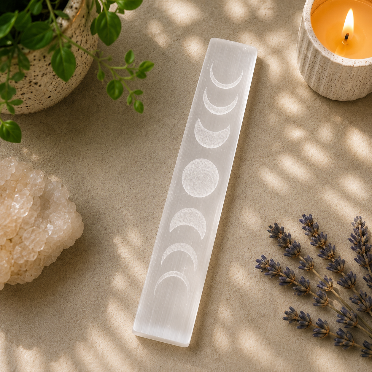 Natural white selenite charging plate engraved with the phases of the moon, displayed on a warm neutral surface with soft natural lighting. The silky translucent crystal features etched lunar phase symbols, while a lit candle, green potted plant, lavender sprigs, and a white quartz cluster create a calming, celestial, zen-inspired aesthetic.