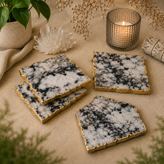 Set of four handcrafted black-and-white natural stone crystal coasters with elegant gold edges displayed on soft beige linen. The coasters are styled beside a lit candle, quartz crystal cluster, white sage bundle, lush greenery, and dried botanicals in a warm, calming boho-inspired setting.