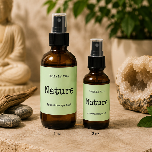 Bella La’ Vita Nature Aromatherapy Mist in 4 oz and 2 oz amber glass spray bottles displayed on a tranquil, spa-inspired backdrop. The soft green labels feature the scent name "Nature" in a minimalist design. The bottles are styled with smooth river stones, natural driftwood, a crystal geode, fresh green leaves, and a softly blurred Buddha statue, evoking the fresh, grounding essence of nature and a peaceful wellness atmosphere.