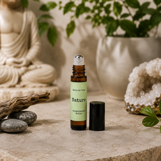 Bella La’ Vita Nature Aromatherapy Roller in an amber glass roll-on bottle with the cap removed to reveal a stainless steel rollerball applicator. The soft green label features the scent name "Nature" in a minimalist design. Displayed on a tranquil, spa-inspired backdrop with smooth river stones, natural driftwood, a sparkling crystal geode, fresh green leaves, and a softly blurred Buddha statue, creating a fresh, grounding atmosphere inspired by the beauty of nature.