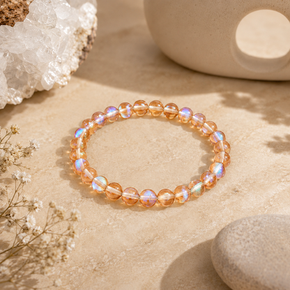Orange Mystic Glass stretch bracelet featuring shimmering translucent orange beads with iridescent flashes of pink, blue, and gold, displayed on a warm stone surface beside clear quartz, dried botanicals, a smooth river stone, and a ceramic wax warmer in a calming, zen-inspired setting.
