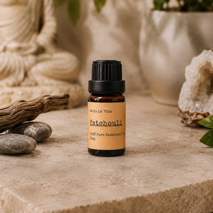 Amber 10 ml bottle of Bella La' Vita Patchouli 100% Pure Essential Oil displayed on a warm zen-inspired stone surface with a Buddha statue, driftwood, smooth stones, greenery, and a crystal geode in the background.