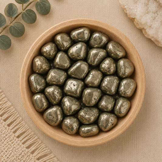 Natural polished pyrite tumbled stones displayed in a round bamboo bowl on a soft beige linen background. The metallic gold gemstones feature a reflective, mirror-like luster with natural mineral flecks and unique shapes. Eucalyptus leaves, a woven cream textile, and a white quartz cluster complete the warm, minimalist, zen-inspired flat lay.