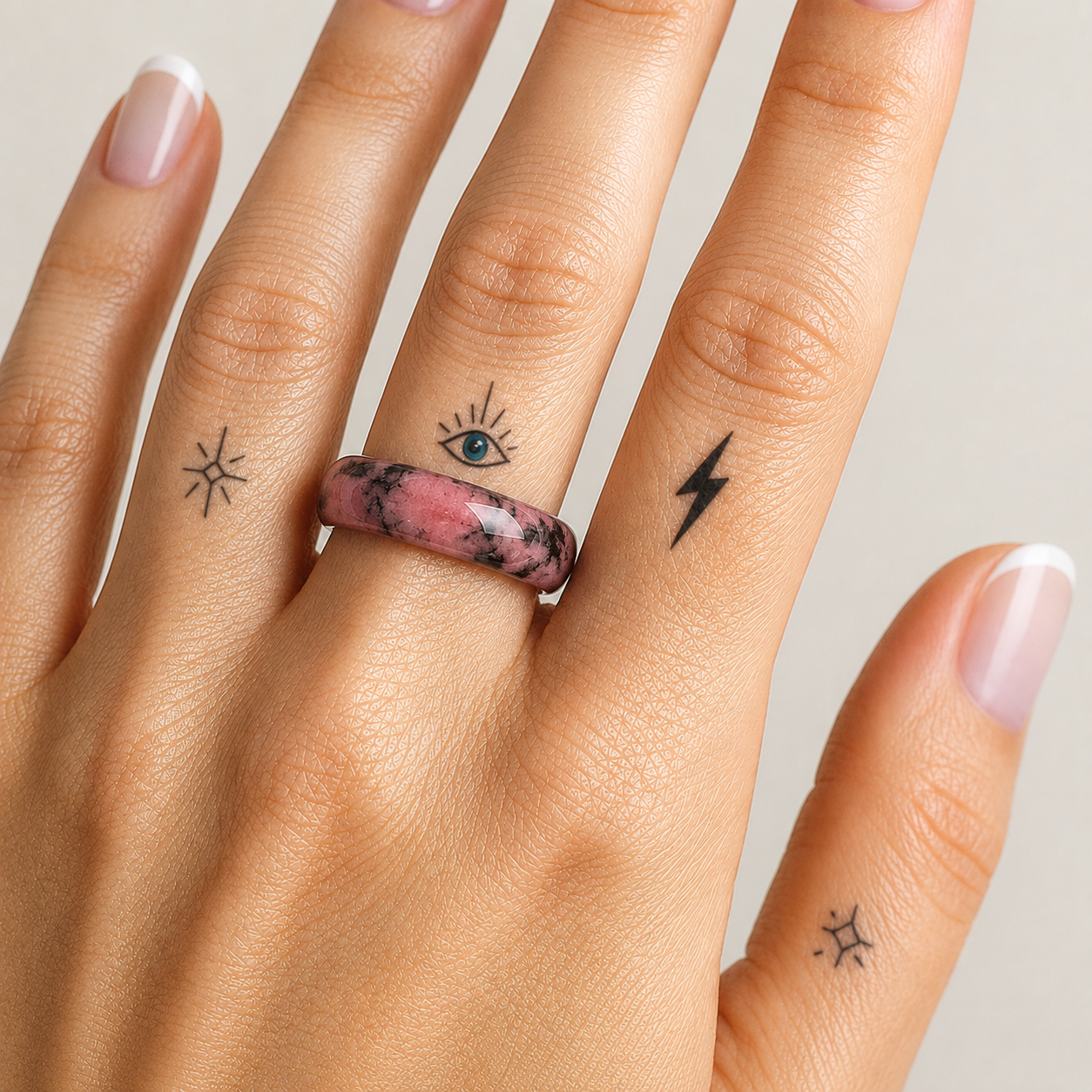 Close-up of a hand wearing a polished rhodonite gemstone ring on the middle finger. The natural stone band features soft pink tones with distinctive black manganese veining, creating a unique marbled pattern. The hand has a French manicure and minimalist finger tattoos, including an eye, lightning bolt, and star designs, set against a soft neutral background with bright natural lighting.