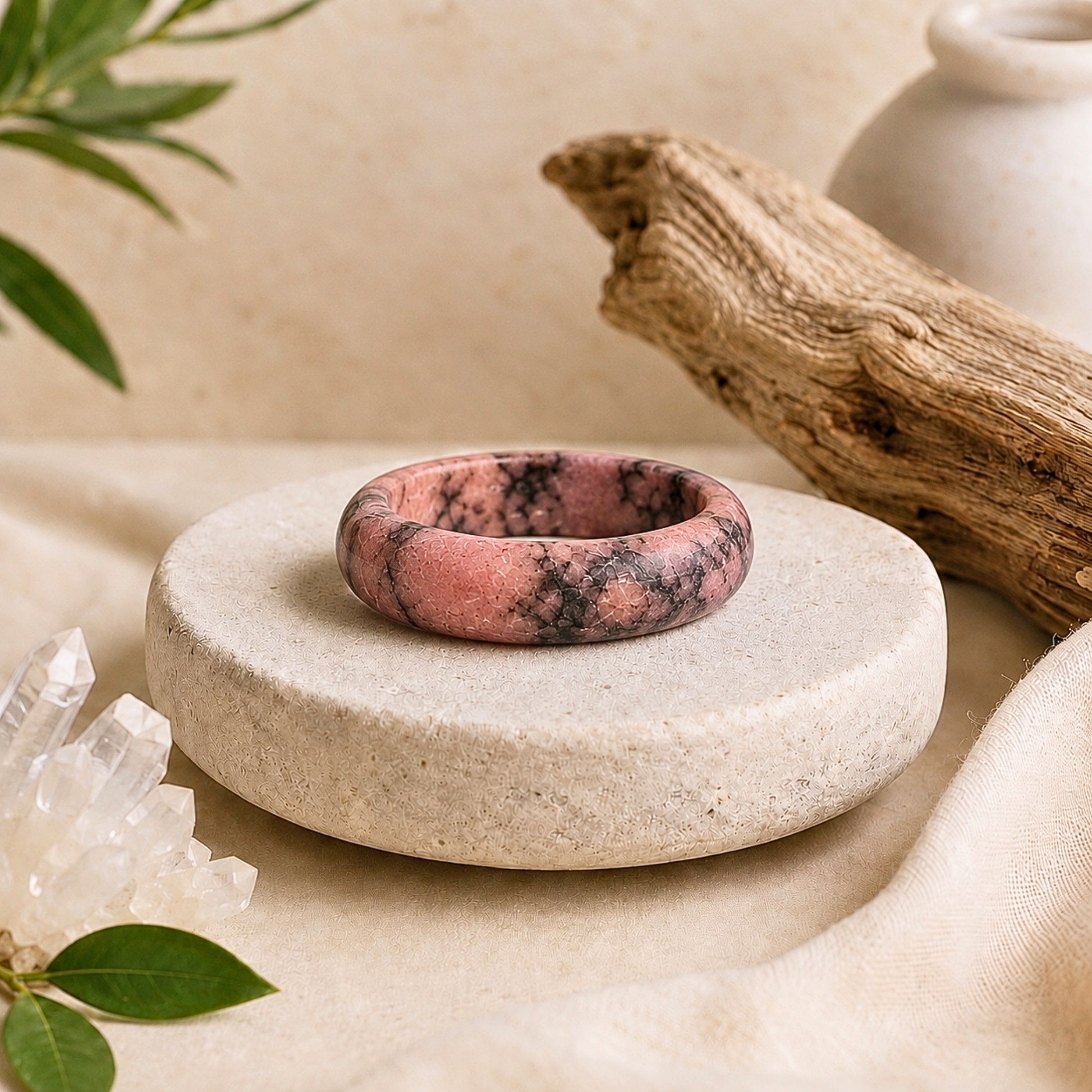 Polished rhodonite crystal ring with natural pink and black marbling displayed on a textured cream stone pedestal, surrounded by clear quartz, driftwood, greenery, and soft beige fabric.