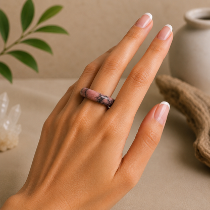 Polished rhodonite crystal ring with natural pink and black marbling worn on a hand with a French manicure, styled against a warm beige zen-inspired background with greenery and driftwood.