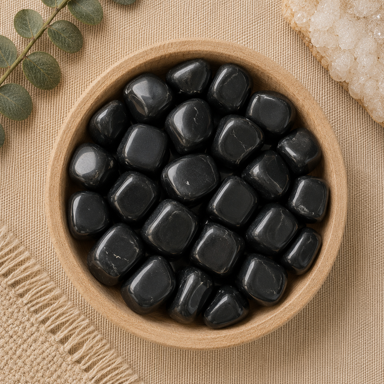 Natural polished black obsidian tumbled stones displayed in a round bamboo bowl on a soft beige linen background. The glossy black gemstones feature smooth, rounded shapes with a deep reflective finish and subtle natural variations. Eucalyptus leaves, a woven cream textile, and a white quartz cluster complete the warm, minimalist, zen-inspired flat lay.