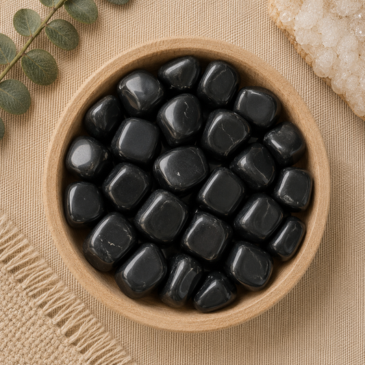 Natural polished black obsidian tumbled stones displayed in a round bamboo bowl on a soft beige linen background. The glossy black gemstones feature smooth, rounded shapes with a deep reflective finish and subtle natural variations. Eucalyptus leaves, a woven cream textile, and a white quartz cluster complete the warm, minimalist, zen-inspired flat lay.