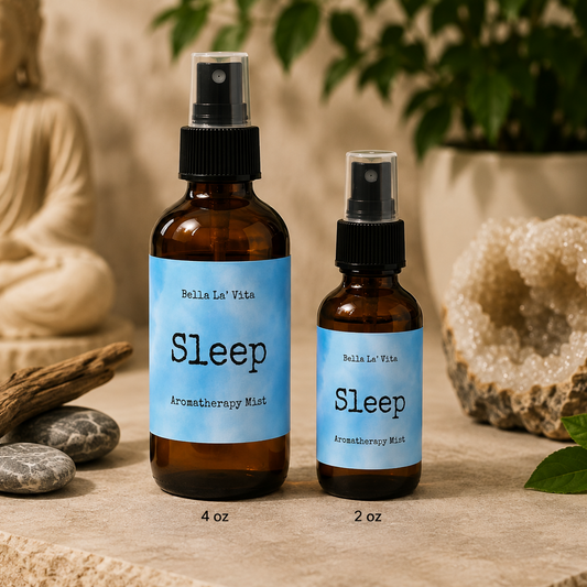 Bella La’ Vita Sleep Aromatherapy Mist in 4 oz and 2 oz amber glass spray bottles displayed on a peaceful, spa-inspired backdrop. The soft blue labels feature the scent name "Sleep" in a minimalist design. The bottles are styled with smooth river stones, natural driftwood, a sparkling crystal geode, fresh green leaves, and a softly blurred Buddha statue, creating a tranquil atmosphere perfect for evening relaxation and winding down.