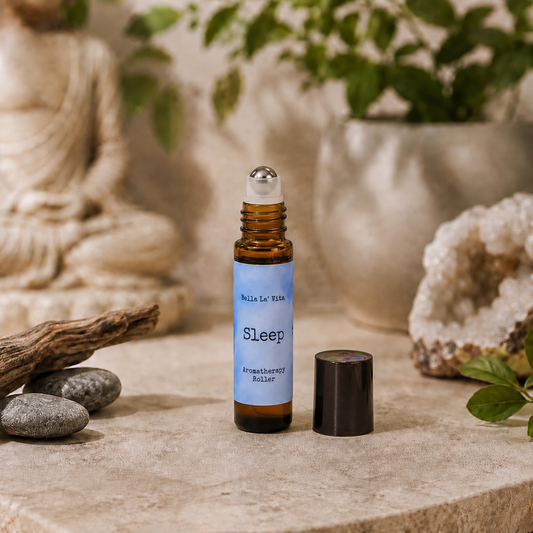 Bella La’ Vita Sleep Aromatherapy Roller in an amber glass roll-on bottle with the cap removed to reveal a stainless steel rollerball applicator. The soft blue label features the scent name "Sleep" in a minimalist design. Displayed on a tranquil, spa-inspired backdrop with smooth river stones, natural driftwood, a sparkling crystal geode, fresh green leaves, and a softly blurred Buddha statue, creating a peaceful atmosphere ideal for relaxation and an evening self-care ritual.