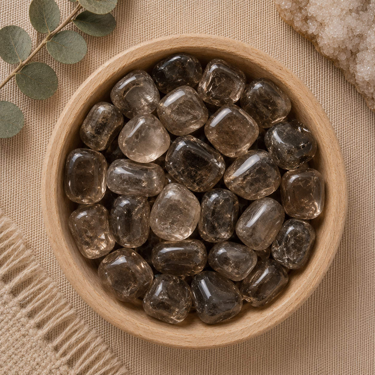 Natural polished smoky quartz tumbled stones displayed in a round bamboo bowl on a soft beige linen background. The translucent gemstones feature rich smoky brown to gray tones with natural internal inclusions and a glossy polished finish. Eucalyptus leaves, a woven cream textile, and a white quartz cluster complete the warm, minimalist, zen-inspired flat lay.