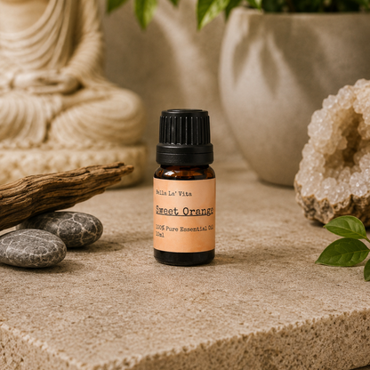 Amber 10 ml bottle of Bella La' Vita Sweet Orange 100% Pure Essential Oil displayed on a warm zen-inspired stone surface with a Buddha statue, driftwood, smooth stones, greenery, and a crystal geode in the background.