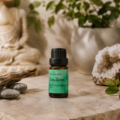 Amber 10 ml bottle of Bella La' Vita Tea Tree 100% Pure Essential Oil displayed on a warm zen-inspired stone surface with a Buddha statue, driftwood, smooth stones, greenery, and a crystal geode in the background.