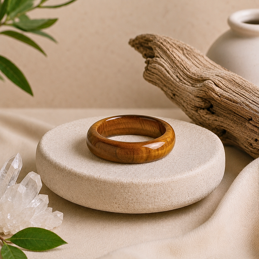 Polished tiger’s eye crystal ring with natural golden-brown banding displayed on a textured cream stone pedestal, surrounded by clear quartz, driftwood, greenery, and soft beige fabric.