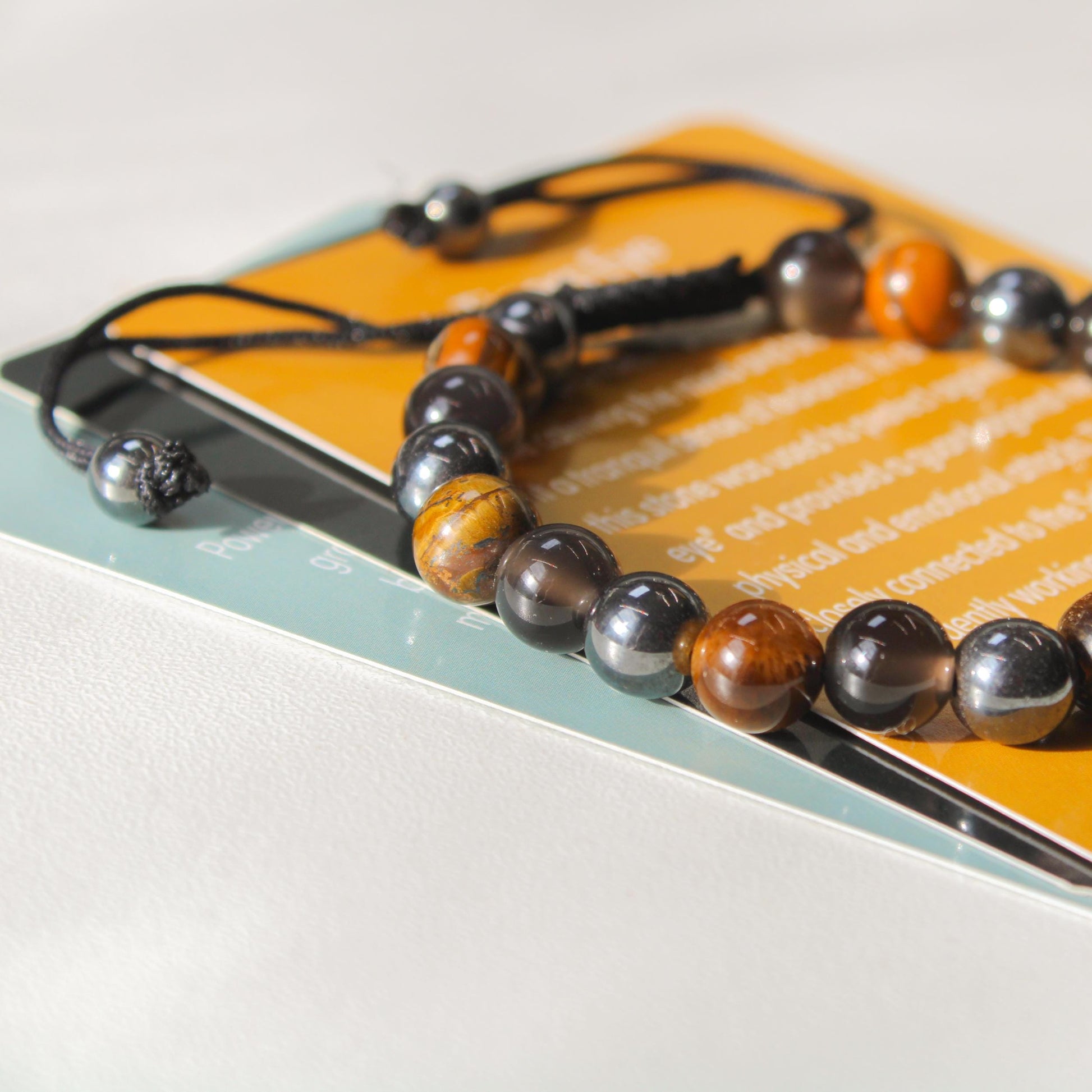 Azenway Triple Protection Bracelet resting on meaning cards, highlighting the mixed Tiger’s Eye, Black Obsidian and Hematite gemstones with the braided adjustable cord ends.