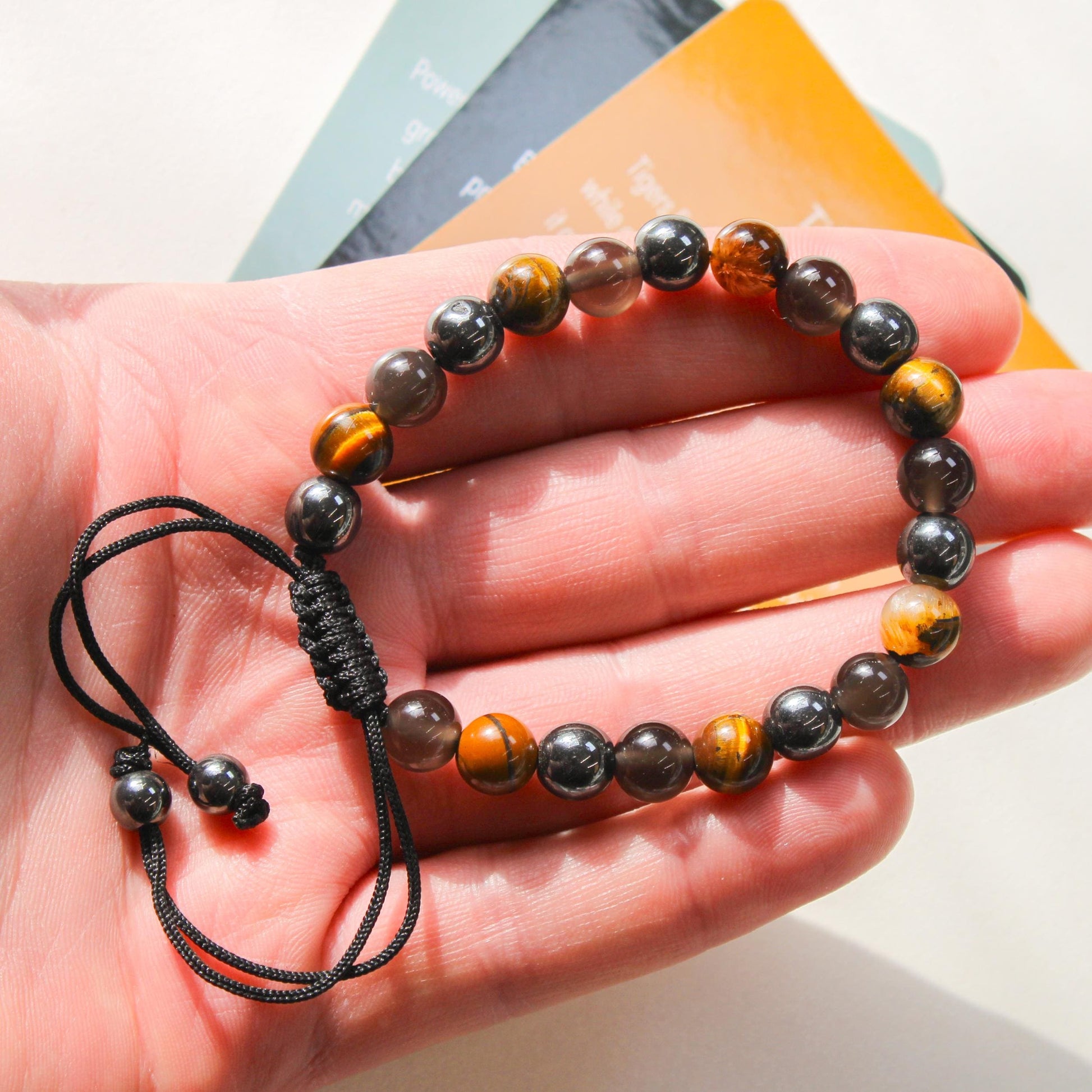 Close-up of the Azenway Triple Protection Bracelet held in the hand, showing polished Tiger’s Eye, Black Obsidian and Hematite beads with a woven adjustable cord.